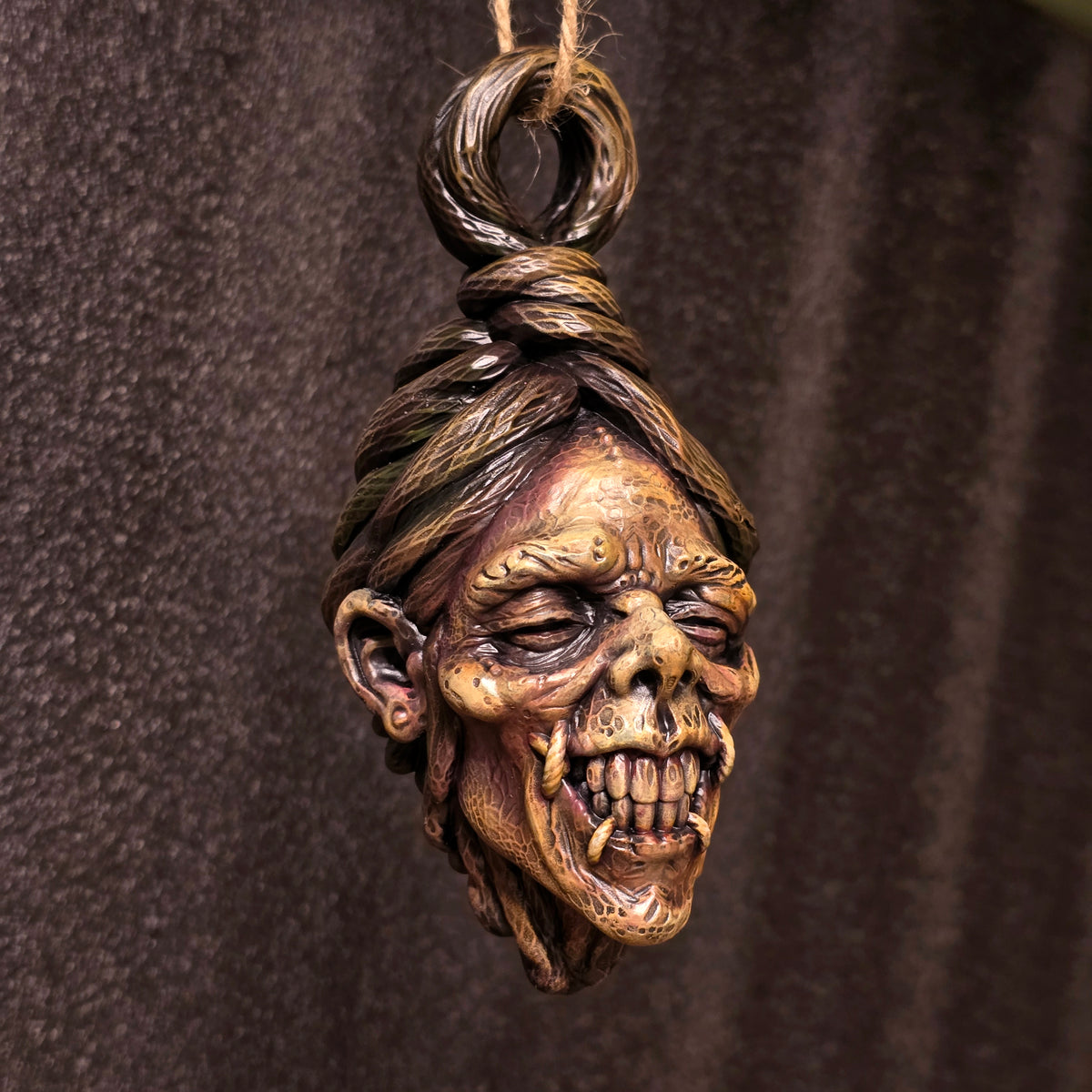 Hand-painted hanging shrunken head sculpture with exposed teeth, deeply creased skin and sculpted hair against a dark background.
