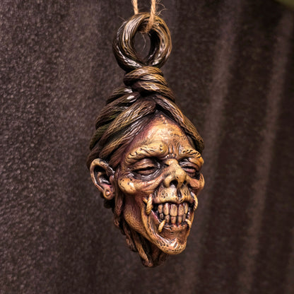 Hand-painted hanging shrunken head sculpture with exposed teeth, deeply creased skin and sculpted hair against a dark background.