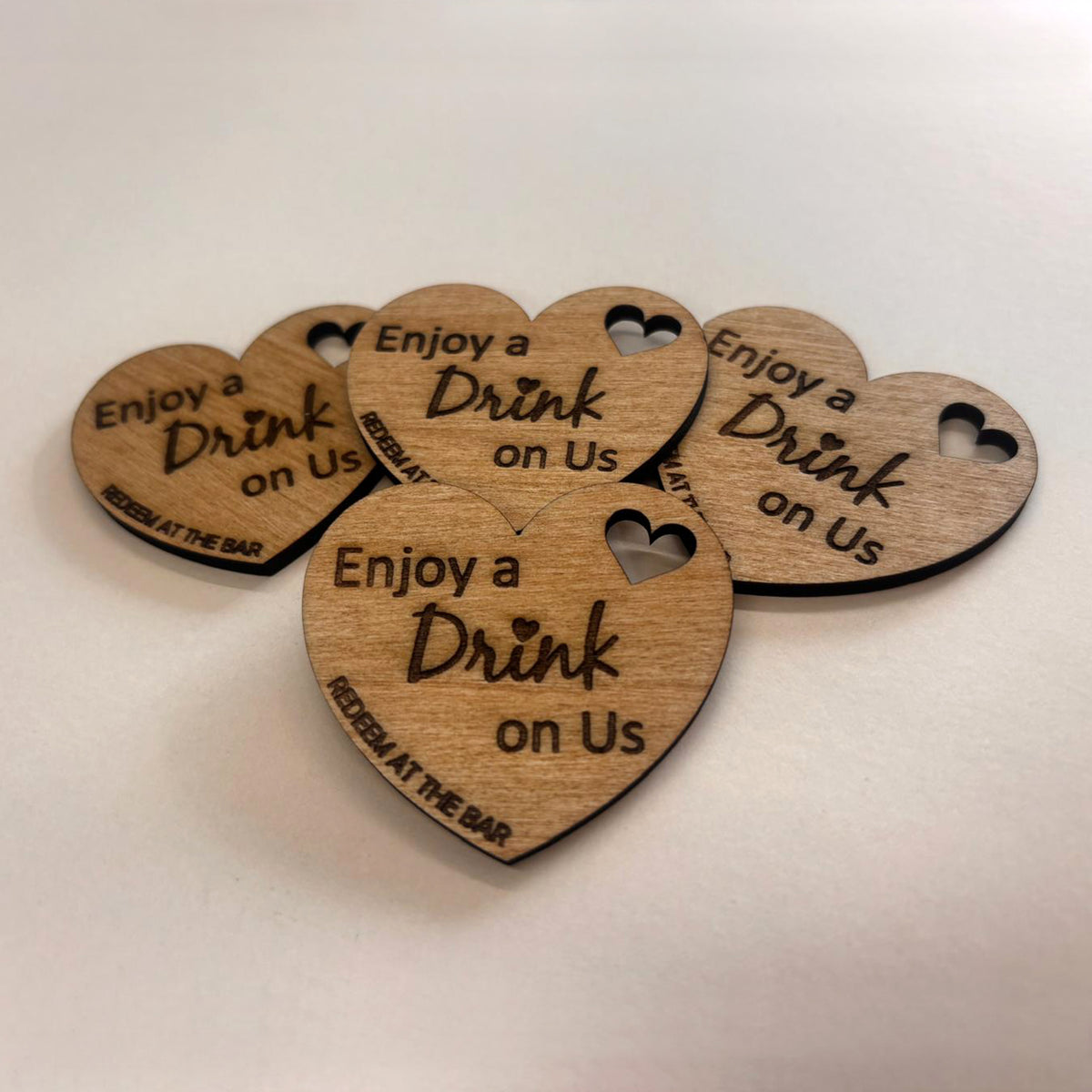Heart-shaped wooden wedding drink tokens engraved Enjoy a Drink on Us, with small heart cut-outs and bar redemption wording.