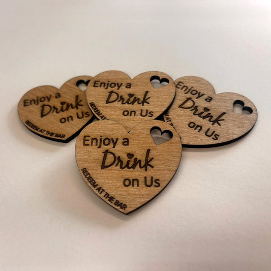Heart-shaped wooden wedding drink tokens engraved Enjoy a Drink on Us, with small heart cut-outs and bar redemption wording.