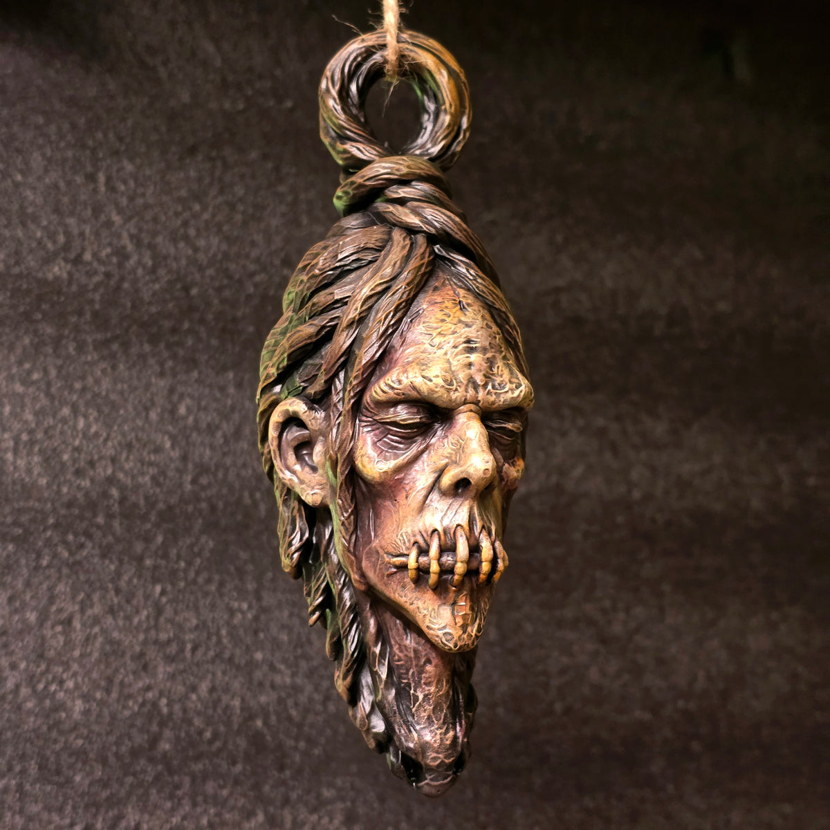 Hanging shrunken head sculpture with stitched mouth, elongated face and weathered hand-painted finish against a dark background.