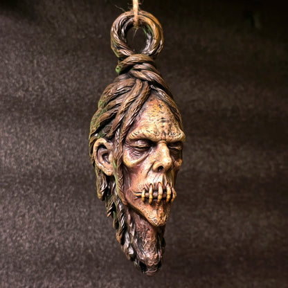 Hanging shrunken head sculpture with stitched mouth, elongated face and weathered hand-painted finish against a dark background.