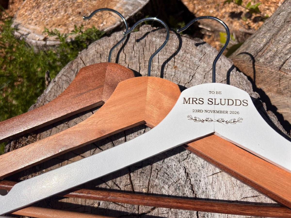 Personalised white engraved bridal hanger with natural light and dark wood colour options on a rustic log.